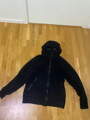 Svart hoodiejacka med goggles C.P company - Svart hoodiejacka från C.P. Company med ikoniska goggles i huvan. Jackan har två fickor med dragkedja och hel dragkedja framtill. Tillverkad i mjuk bomull, perfekt för dig som vill sticka ut med streetwear-vibe. Snygg och funktionell design. Jag är 170-173 och väger 53 kg och den passar bra på mig. Ändå om de är storlek L så passar den mig och jag är en S/M person.