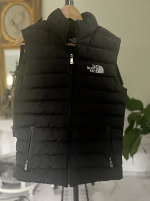 Svart dunväst från The North Face - Svart dunväst från The North Face Summit Series med quiltad design och logga på bröstet. Västen har två praktiska fickor med dragkedja och hög krage. Perfekt för lager-på-lager och outdoor vibes.