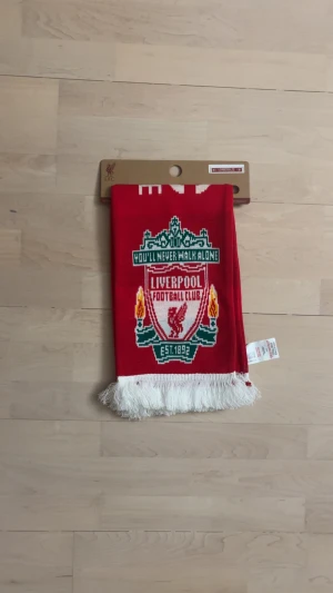 Liverpool FC röd halsduk med tofsar - En helt ny röd halsduk från Liverpool FC med klubbens klassiska logga och texten 'You'll Never Walk Alone' i grönt och vitt. Halsduken har vita tofsar i ändarna och är perfekt för att visa ditt stöd på matchen. Tjockt stickat material som håller dig varm.