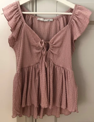 Rosa blus med volang och snörning - Supersöt rosa blus från 157 med volangdetaljer på axlarna och v-ringning med snörning framtill. Blusen har en luftig, crinklad struktur och är lätt utsvängd nedtill för en flowy look. Perfekt för dig som gillar romantisk stil och mjuka färger.