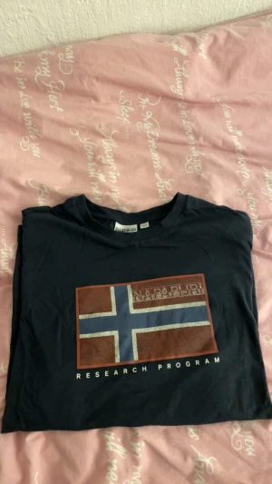 Svart t-shirt från Napapijri - Snygg svart t-shirt från Napapijri med tryck av norsk flagga och texten 'Research Program' på bröstet. Klassisk rund hals och korta ärmar. Perfekt för dig som gillar streetwear och vill ha en stilren look.
