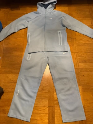 Nike NOCTA ljusblå mjukisbyxor - Snygga ljusblå mjukisbyxor från Nike NOCTA med elastisk midja och snörning. Byxorna har raka ben, dragkedja på ena benet och diskret NOCTA-logga. Materialet är mjukt och skönt, perfekt för chill eller träning. Ikonisk Swoosh på framsidan.