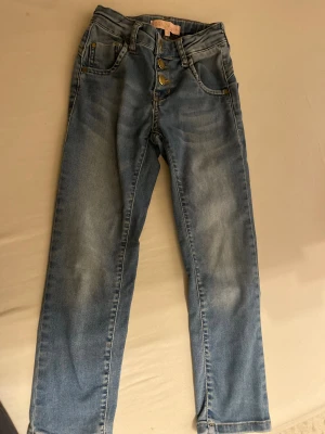 Blå jeansbyxor från Denim, strl 116 - Snygga blå jeansbyxor från Denim by S. I storlek 116. Klassisk femficksmodell med guldfärgade knappar. Hög midja och raka ben, perfekt för dig som gillar en enkel och stilren look. Barn till tjejer 