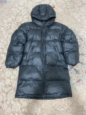 Svart lång pufferjacka från Nike - Svart pufferjacka från Nike med Therma-Fit teknologi, huva och hel dragkedja framtill. Jackan är lång och har en clean look med diskret Nike-logga på bröstet. Perfekt för kalla dagar när du vill hålla värmen och ändå se chill ut.