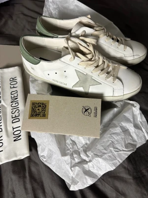 Golden Goose Superstar sneakers vit/grön - Golden Goose Superstar sneakers i vitt skinn med ljusgrön häl och ikonisk stjärndetalj i grå mocka på sidan. Beige snörning och platt sula. Snygg och clean design med coola vintage-vibbar, perfekt för dig som gillar streetstyle.