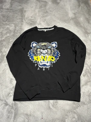 Svart Kenzo sweatshirt med tigerbroderi  - Svart sweatshirt från Kenzo i storlek XL med ikoniskt broderat tigerhuvud i blått, vitt, gult och silver på bröstet. Kenzo-loggan i gult över motivet och Paris-broderi under. Tröjan har rund hals och långa ärmar, tillverkad i mjuk bomull.