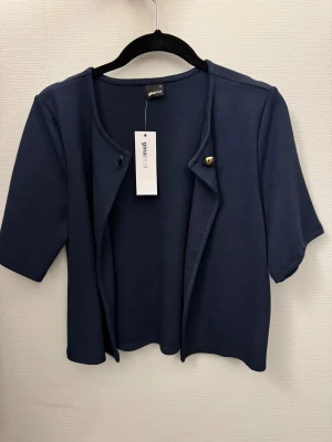 Mörkblå kofta från Gina Tricot - Snygg mörkblå kofta från Gina Tricot med korta ärmar och rund hals. Koftan har en enkel design med guldfärgad knapp upptill och är tillverkad i mjuk bomull. Perfekt att slänga över en topp för en chill look.