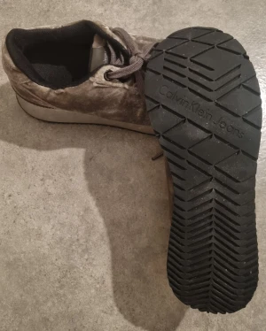 Grå sneakers från Calvin Klein Jeans - Snygga grå sneakers från Calvin Klein Jeans med platt sula och rund tå. Skorna har snörning och är tillverkade i ett mjukt, sammet-liknande material som ger en lyxig känsla. Perfekta för dig som gillar stilrena och moderna skor.