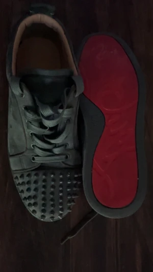 Mörkgrå mocka sneakers med nitar - Säljer ett par mörkgrå sneakers i mocka med coola nitar på tån och snörning framtill. Insidan är beige och sulan har en röd undersida som sticker ut. Skorna har rund tå och platt sula, perfekt för dig som gillar edgy detaljer.