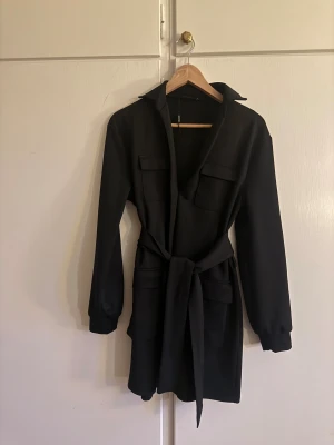 Svart kort klänning med bälte - Stilren svart klänning med lång ärm, djup v-ringning och knytbälte i midjan. Klänningen har stora fickor framtill och en klassisk krage som ger en cool look. Perfekt för dig som gillar minimalistisk stil och vill ha något enkelt men snyggt.