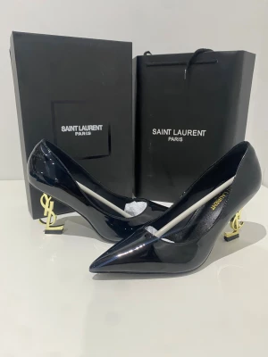 Svarta Saint Laurent pumps med YSL-klack - Säljer ett par svarta pumps från Saint Laurent med glansig finish och spetsig tå. Klacken är i guld och formad som YSL-loggan, vilket ger en riktigt lyxig vibe. Skorna är tillverkade i skinn och har en elegant, modern design som sticker ut.