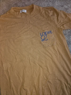 Beige t-shirt med broderad ficka - Snygg beige t-shirt i bomull med rund hals och korta ärmar. På bröstet finns en broderad ficka med Loewe-loggan i blått och beige, vilket ger en unik och cool detalj. Perfekt för dig som gillar stilrena plagg med exklusiv känsla.