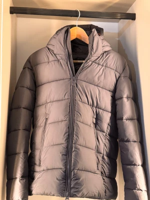  Save The Duck  - Säljer en grå pufferjacka från Save The Duck i storlek XS. Jackan har huva, två fickor med dragkedja och är tillverkad i återvunnen polyester med Plumtech-fyllning. Snygg, stilren och perfekt för kyliga dagar.