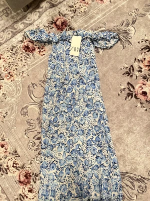 Blommig offshoulder klänning från Zara - Supersöt offshoulder klänning från Zara i vitt och blått med blommigt mönster. Smockad upptill och volangdetalj vid axlarna. Perfekt för sommarens alla tillfällen. Lätt och luftig känsla. Helt ny