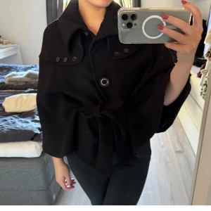 Poncho Jacka - Snygg svart kappa med stor krage, dekorativa knappar upptill och knytband i midjan. Modellen är loose och har vida ärmar som ger en cool och avslappnad look. Perfekt för dig som vill ha en stilren och trendig jacka till höst och vinter.