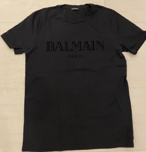 Svart Balmain Paris t-shirt - Snygg orginal svart t-shirt från Balmain Paris med stor logga framtill. Klassisk rund halsringning och korta ärmar. Perfekt för dig som gillar stilrena och exklusiva plagg med statement-tryck.