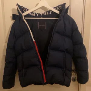 Mörkblå dunjacka Tommy Hilfiger - Snygg mörkblå dunjacka från Tommy Hilfiger med puffad design och logotyp på kragen. Jackan har dragkedja framtill, långa ärmar och detaljer i rött och vitt. Fylld med dun och fjäder för extra värme, perfekt för kalla dagar. Ifall ni vill ha fler bilder kom gärna privat. Kan alltid diskutera ett lägre pris. Storleken är 164