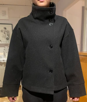 Svart vår jacka med hög krage - Stilren svart skepparkavaj med hög krage och stora knappar framtill. Jackan har en loose passform, raka axlar och långa ärmar med breda muddar. Materialet är tunt och passar därför som en vårjacka. Kragen kan bäras både uppfälld och nedvikt för olika looks.