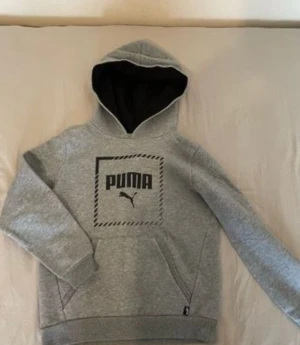 Grå hoodie från Puma  - Säljer en grå hoodie från Puma med svart logga och tryck på bröstet. Tröjan har huva med svart insida, långa ärmar och en stor magficka. Materialet är mjukt och består av bomull och polyester, perfekt för chill eller träning.