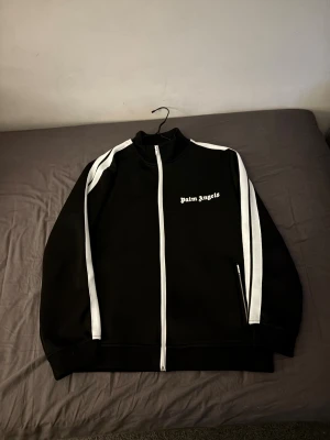 Svart Palm Angels - Palm Angels svart vindjacka med vita ränder längs ärmarna och vit dragkedja. Jackan har ståkrage, två fickor med dragkedja och logotypen tryckt på bröstet. Snygg och sportig design som sticker ut med kontrastdetaljer.