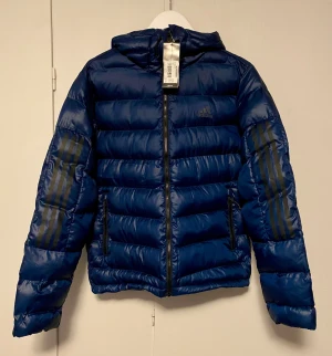 Ny marinblå pufferjacka från Adidas S - Helt ny jacka med tag kvar från Adidas med huva och dragkedja. Jackan har de klassiska tre ränderna på ärmarna och Adidas-logga på bröstet. Tillverkad i glansig polyester och har två sidofickor med dragkedja. Perfekt för kalla dagar. Modell: Itavic 3S