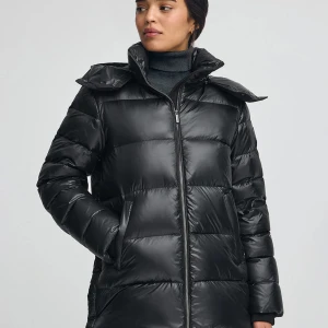 Svart pufferjacka med huva - Svart pufferjacka med hög krage och stor huva. Jackan har quiltad design, dragkedja framtill och två sidofickor. Materialet är glansigt och syntetiskt, vilket ger en modern vibe. Perfekt för kalla dagar och har en loose passform som gör den extra bekväm.