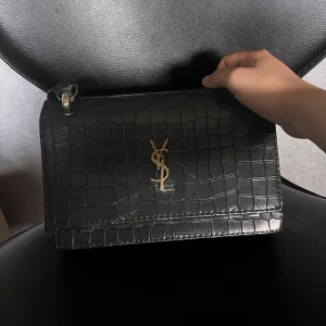 Svart axelväska från  - Säljer en svart axelväska (inte äkta!),  med präglat mönster och guldfärgad logga framtill. Den är lite skadad kolla bilden, Väskan har en stilren design och justerbar axelrem. Perfekt för dig som vill ha en fin accessoar till din outfit.