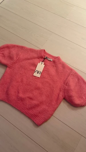 Rosa stickad tröja från ZARA - Superfin rosa stickad tröja från ZARA med puffiga korta ärmar och ribbade muddar. Tröjan har en croppad passform och rund halsringning, perfekt för att lyfta din outfit med en poppig färg.