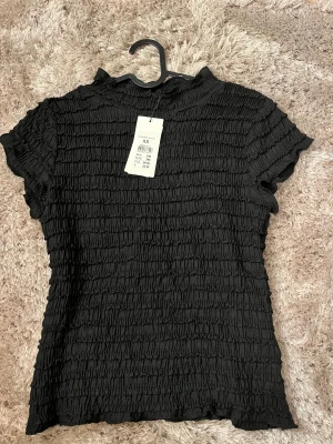Svart ribbad topp XS från Vero Moda - Säljer en svart ribbad topp från Vero Moda i storlek XS. Toppen har korta ärmar, hög hals och ett coolt rynkat tyg som ger en unik struktur. Perfekt för dig som gillar stilrena och trendiga plagg med lite edge.