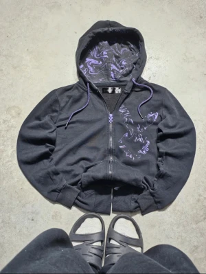 Svart playboy hoodie med lila mönster - Svart hoodie med dragkedja och huva, dekorerad med lila swirl-mönster på bröstet och insidan av huvan. Lila snören och ribbade muddar vid ärmslut och nederkant. 