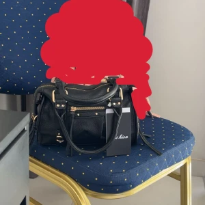 Svart handväska med gulddetaljer - Snygg svart handväska i skinnimitation med guldfärgade metalldetaljer och dragkedjor. Väskan har både handtag och axelrem, samt flera fack med dragkedja framtill. Perfekt storlek för att få plats med allt du behöver och ger en cool vibe till din outfit.