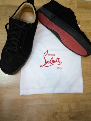 Christian Louboutin svarta sneakers - Svarta sneakers från Christian Louboutin med klassisk röd sula och signatur på undersidan. Skorna är tillverkade i mocka med detaljer i skinn. Snygg och stilren design som sticker ut med den ikoniska röda sulan. Satt nyss in helt nya skosnören och ända defekten är att skorna missar en knapp på toppen fast det är inget som syns!