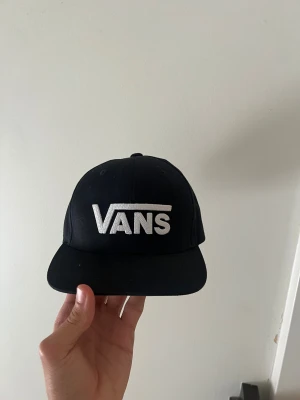 Svart Vans snapback keps - Snygg svart snapback keps från Vans med vit broderad logga framtill. Klassisk platt skärm och justerbar plastrem bak för perfekt passform. Kepsen är tillverkad i bomull och har en stilren, urban look som passar till streetwear.