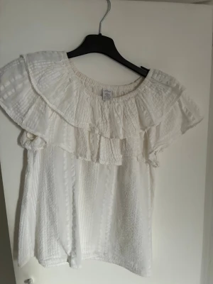 Vit blus med volang från Lindex - Säljer en vit blus från Lindex med breda volanger över axlarna och runt halsen. Även om den är i barnstorlek skulle jag säga att den passar dig som har XS!