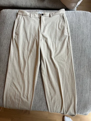 Beiga kostymbyxor Jack & Jones 32/32 - Snygga beiga kostymbyxor från Jack & Jones Pants Studio i relaxed fit. Byxorna har raka ben, bälteshällor, sidofickor och bakfickor med knapp. Perfekta för en clean och stilren look. Materialet är mjukt och känns bekvämt mot huden.