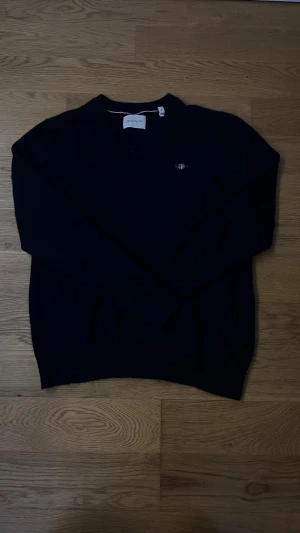 Mörkblå stickad tröja från GANT - Snygg mörkblå stickad tröja från GANT med v-ringad hals och diskret logga broderad på bröstet. Tröjan har långa ärmar och klassisk passform, perfekt för dig som gillar stilrena och tidlösa plagg. Materialet är mjukt och skönt mot huden.