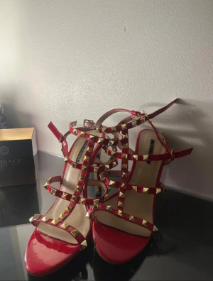 Rockstud röda sandaler - Säljer ett par röda  Rockstud sandaler med hög klack och öppen tå. Skorna har flera remmar dekorerade med guldiga nitar och en snygg, glansig finish. Perfekt för dig som vill sticka ut med en edgy och lyxig look. F🅰️ǩr