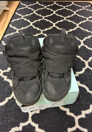 Svarta Lanvin sneakers med breda snören - Lanvin sneakers i helsvart med breda, mönstrade snören och rund tå. Snyggt designade med detaljer i skinn och mesh, samt metallöglor för snörningen. Kommer med originalkartong. Perfekta för dig som gillar chunky och stilrena sneakers.