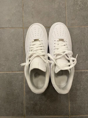 Nike Air Force 1 vita sneakers - Fräscha Nike Air Force 1 sneakers i helvitt med klassisk låg siluett. Skorna har perforerad tå, snörning och platt sula. Tillverkade i skinn med mjuk textil på insidan. Swoosh-logga på sidan och metallplatta vid snörningen för extra stilpoäng.