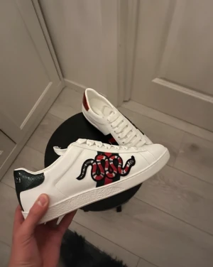 Gucci Ace sneakers med orm - Gucci Ace sneakers i vitt skinn med ikonisk röd och svart orm-brodyr på sidan, grönt och rött metallic-hälparti med Gucci-logga. Klassiska gröna och röda ränder på sidan, platt sula och snörning. Perfekta för dig som vill sticka ut med lyxig streetstyle.