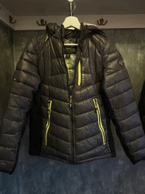 Svart pufferjacka med gula detaljer - Snygg svart pufferjacka från Geographical Norway med huva och coola gula dragkedjor. Jackan har quiltad design, lång ärm och logga på ärmen samt ryggen. Perfekt för dig som gillar sportig stil och vill sticka ut med färgdetaljer.