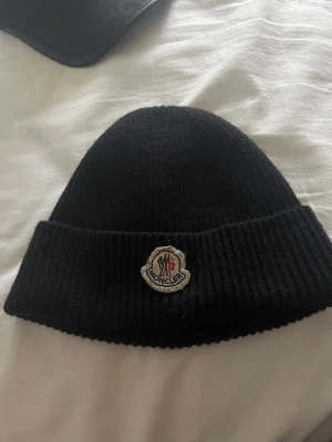 Svart Moncler mössa i ribbstickad ull - Svart ribbstickad mössa från Moncler med klassisk uppvikt kant och broderad Moncler-logga framtill. Snygg och enkel design som passar perfekt när det är kallt ute. Materialet är mjuk ull som håller dig varm och loggan ger en exklusiv touch.