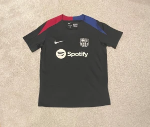 FC Barcelona svart Nike fotbollströja  - Säljer en svart FC Barcelona fotbollströja från Nike med Spotify-logga på bröstet. Tröjan har korta ärmar och detaljer i rött och blått på axlarna. Materialet är lätt och andas, perfekt för match eller träning. Klassisk klubbsköld på bröstet.