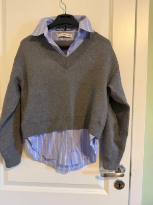 Grå tröja med skjortdetalj från Mango - Snygg grå tröja från Mango med v-ringad hals och långärmad design. Underdelen har en blå skjortdetalj med vita och röda ränder som ger en cool lager-på-lager look.