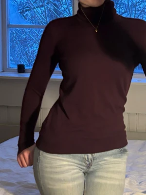 Vinröd polotröja från Ralph Lauren - Snygg vinröd/mörklila polotröja från Lauren Ralph Lauren i slim passform. Tröjan har lång ärm och hög polokrage, perfekt för lager på lager. Materialet är mjukt och stretchigt, vilket gör den skön att bära under kyliga dagar. Nopprig
