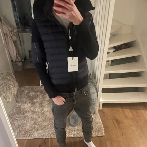Mörkblå moncler cardigan - Snygg mörkblå moncler cardigan med stickade ärmar och quiltad framsida. Västen har en stor huva, dragkedja framtill, ribbade muddar och Moncler-logga på ärmen. Perfekt för lager-på-lager och streetstyle. Loggan skannar. Säljs på grund av att den var för liten 