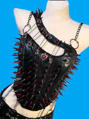 Svart korsett med kedjor och spikes - Unik svart korsett i syntetmaterial med en axel, dekorerad med röda och silvriga spikes, stora metallringar och kedjor. Framsidan har detaljer som hjärtberlock och snörning baktill för justerbar passform. Perfekt för dig som gillar edgy och alternativ stil.