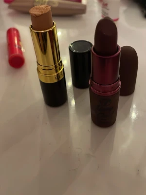 Concealer och mörkt läppstift - Säljer en Revlon concealer i stickformat med guld och svart hylsa, perfekt för att täcka ojämnheter. Medföljer ett mörkbrunt läppstift från W7 i snygg matt brun hylsa med metallisk finish. Båda är smidiga att ha med i väskan.