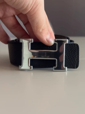 Herme’s bälte  - Bilden visar ett svart bälte med ett stort H-format spänne i silverfärg, inte en väst.helt ny 