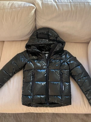 Vinterjacka från Khujo XS - Säljer en glansig mörkgrön pufferjacka från Khujo i storlek XS. Jackan har huva med snörning, två vertikala dragkedjefickor framtill och en markerad quiltad design. Perfekt för kalla dagar och har en modern, boxig passform. Använd fåtal gånger, köpt för 1800kr. 